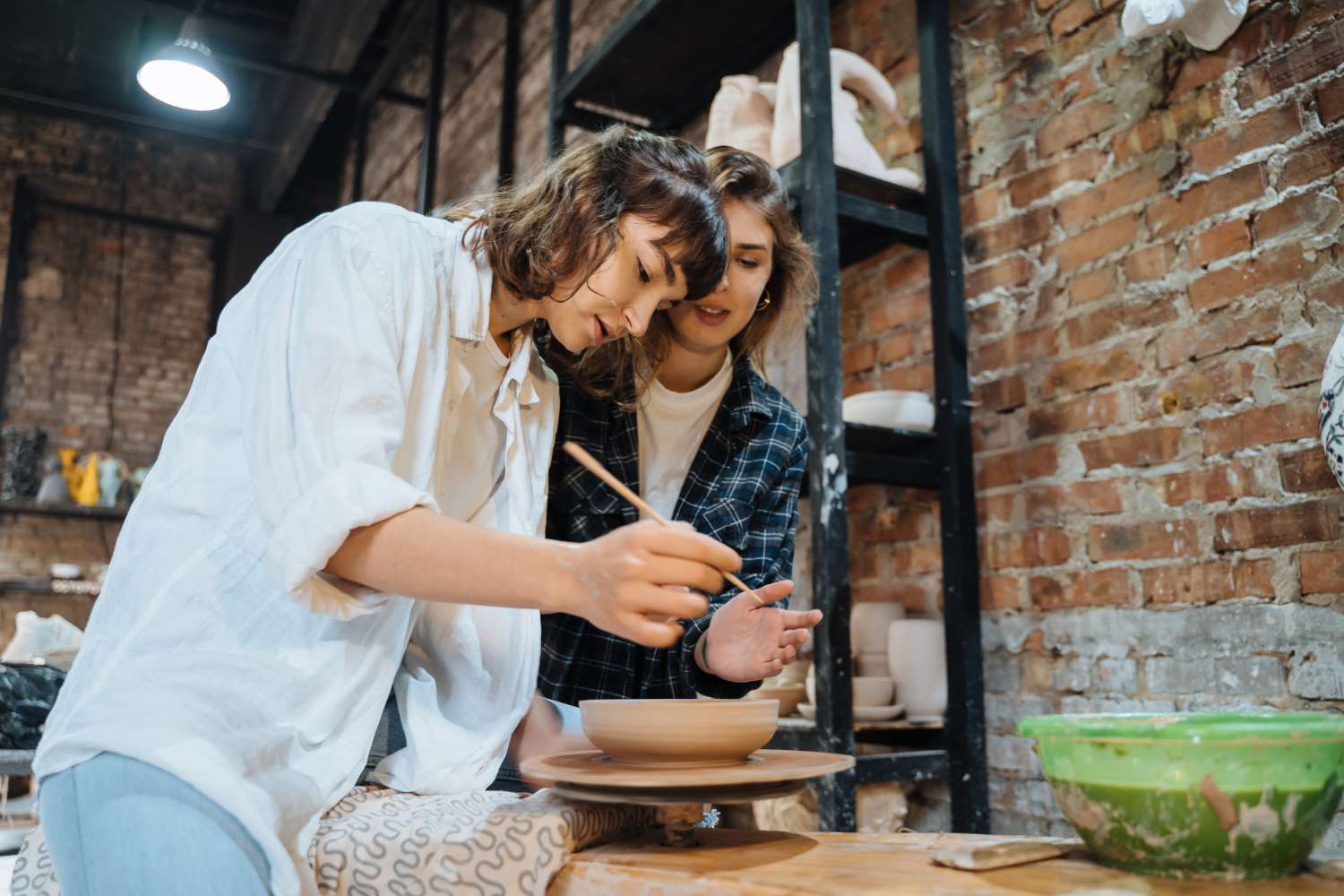 Two female artists working together at a pottery workbench in a bright, brick-walled studio.
