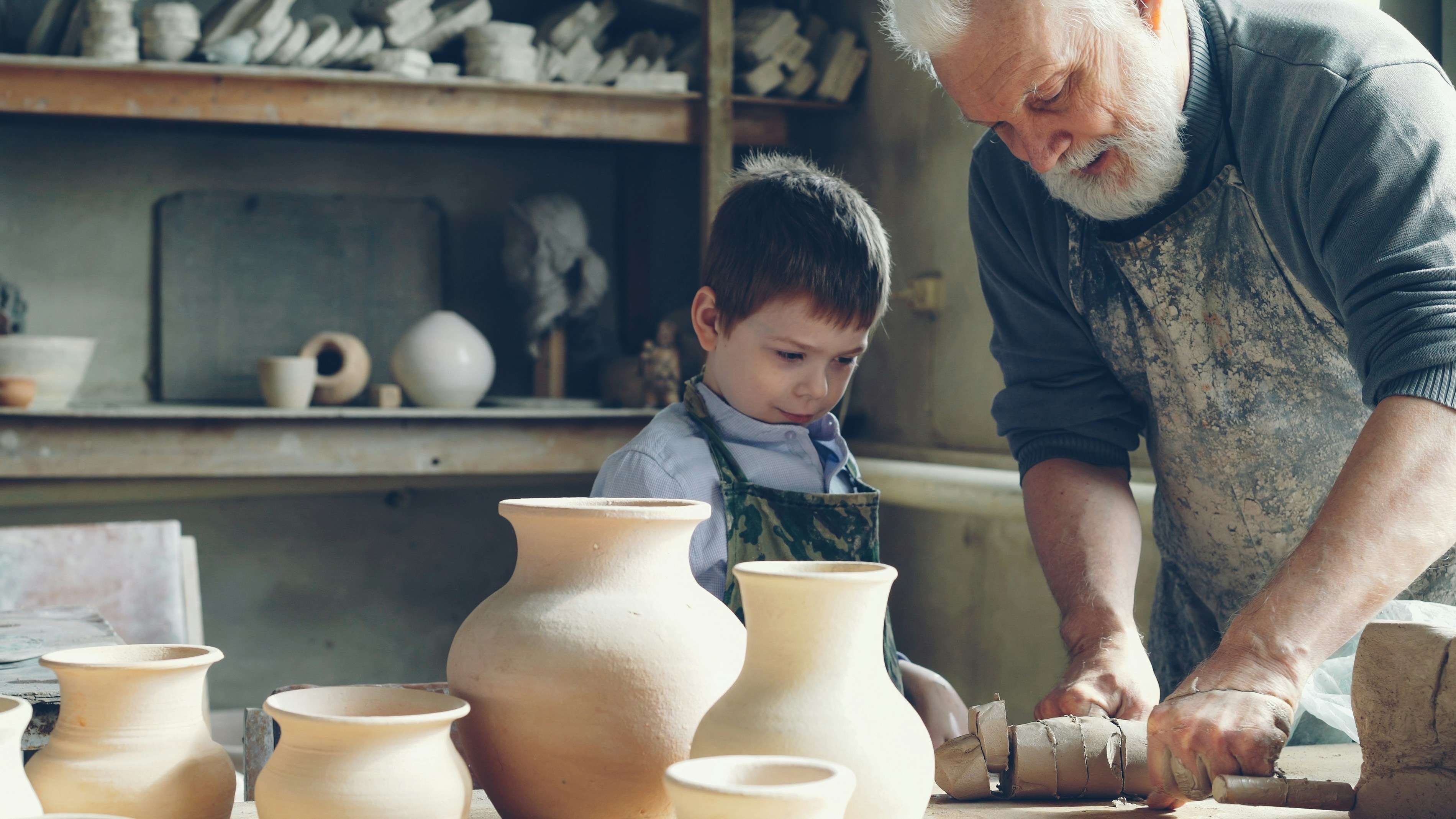 An older man and young boy working side by side in a pottery studio surrounded by ceramic vases.